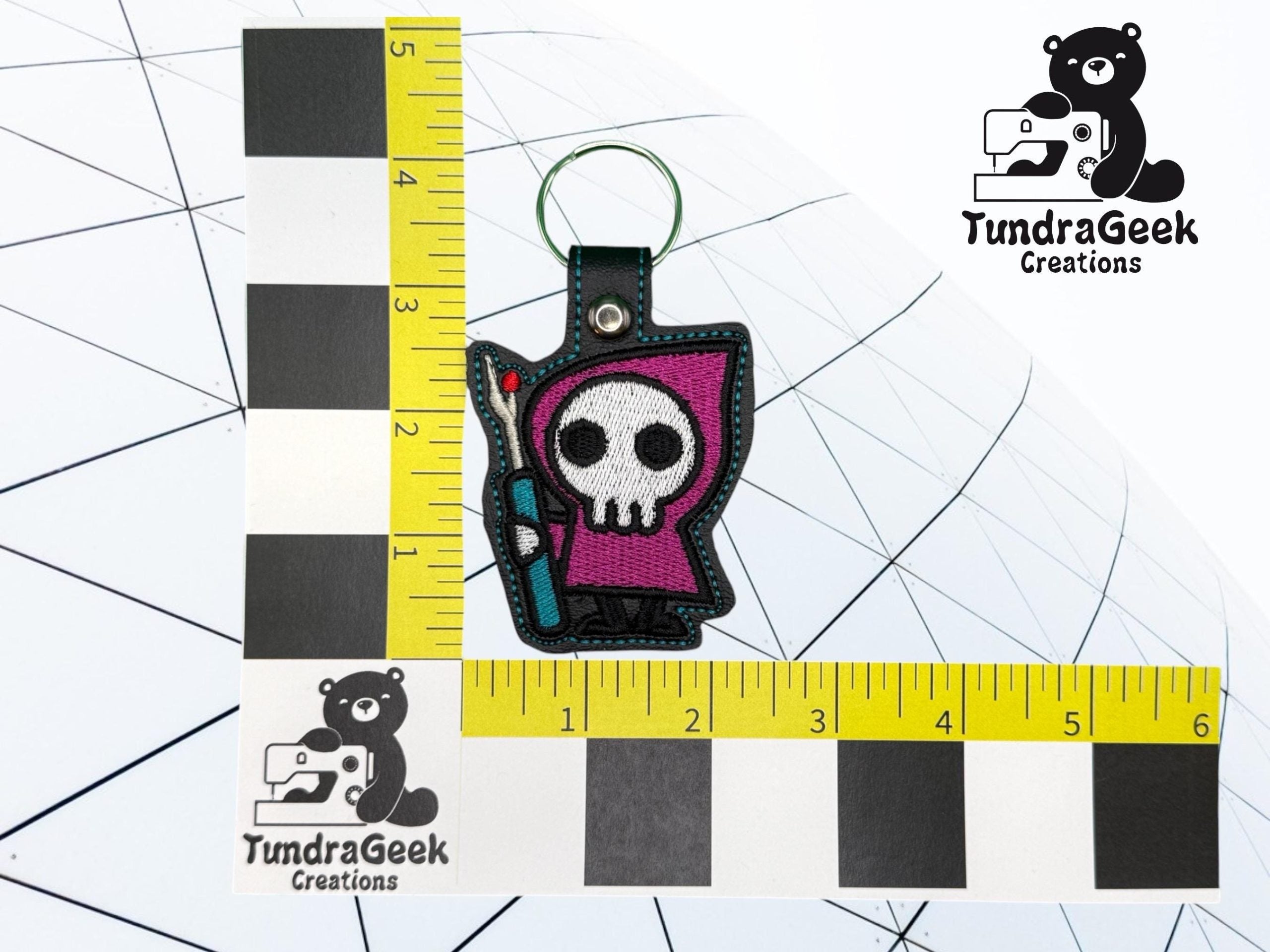 A small keychain with a cartoon grim reaper design is placed on a measurement grid next to the TundraGeek Creations logo.