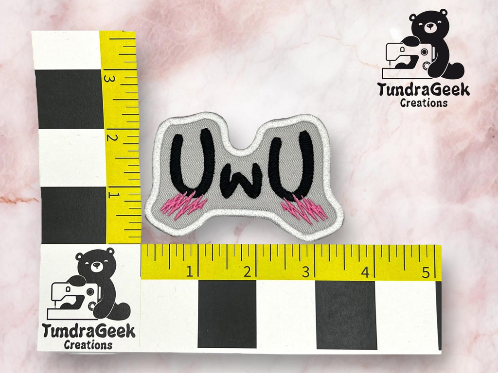 A gray embroidered patch with UWU in black letters and pink accents, placed on a marble background next to measuring tapes indicating size of about 3 inches wide.