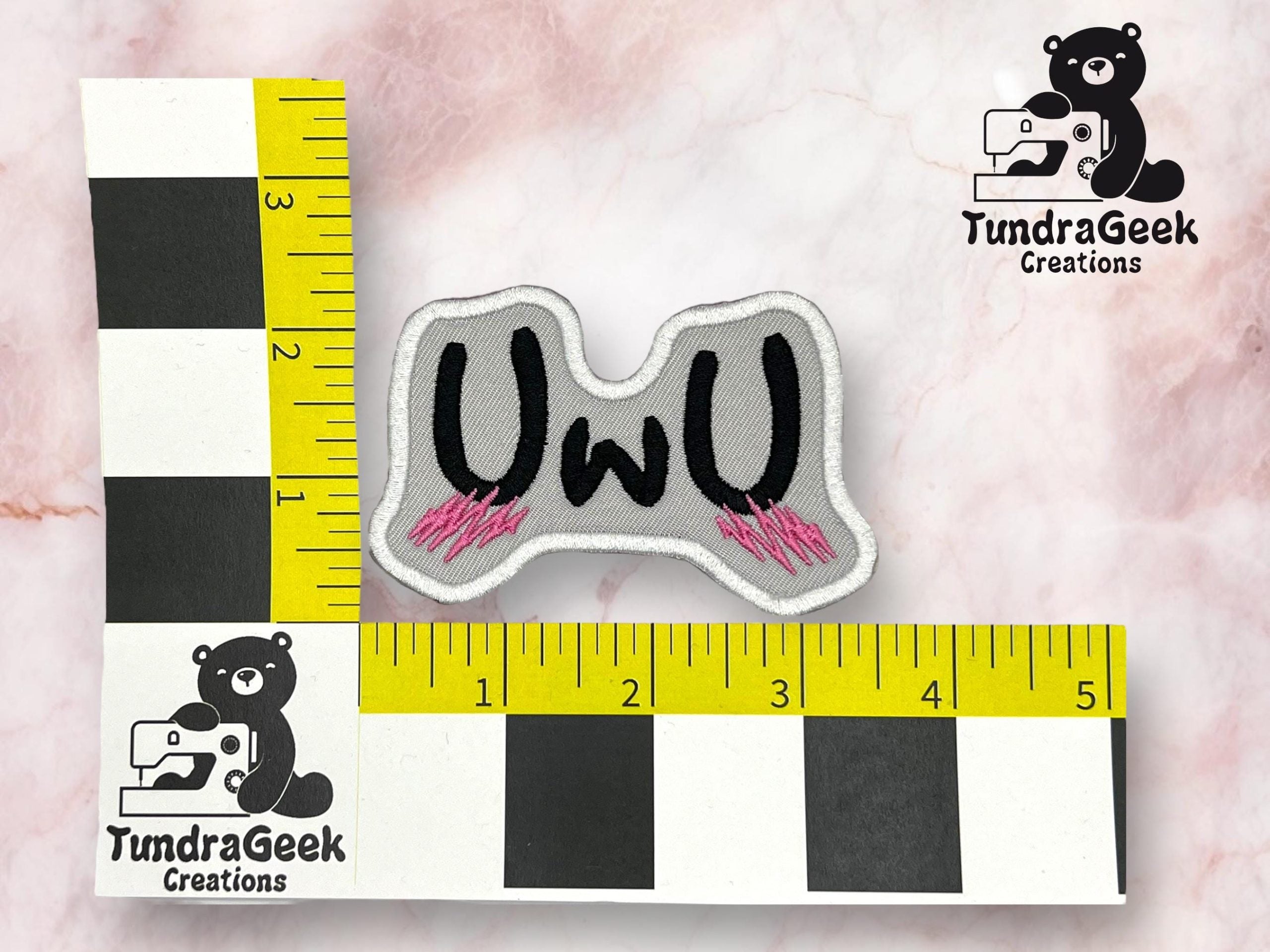 A gray embroidered patch with UWU in black letters and pink accents, placed on a marble background next to measuring tapes indicating size of about 3 inches wide.