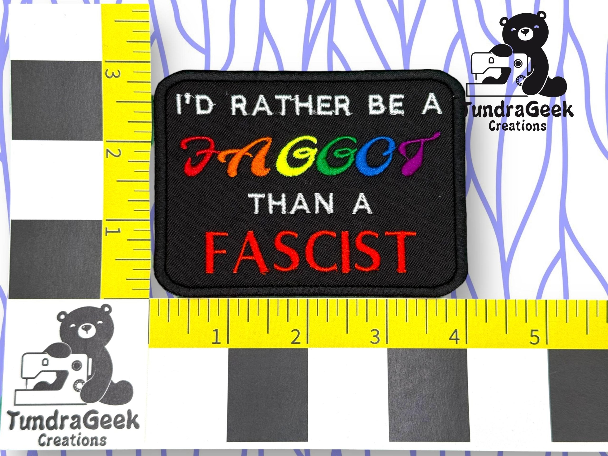 A rectangular patch with the text ID RATHER BE A FAGGOT THAN A FASCIST, with FAGGOT in rainbow colors and FASCIST in red. Yellow rulers and TundaGeek Creations logo surround it.