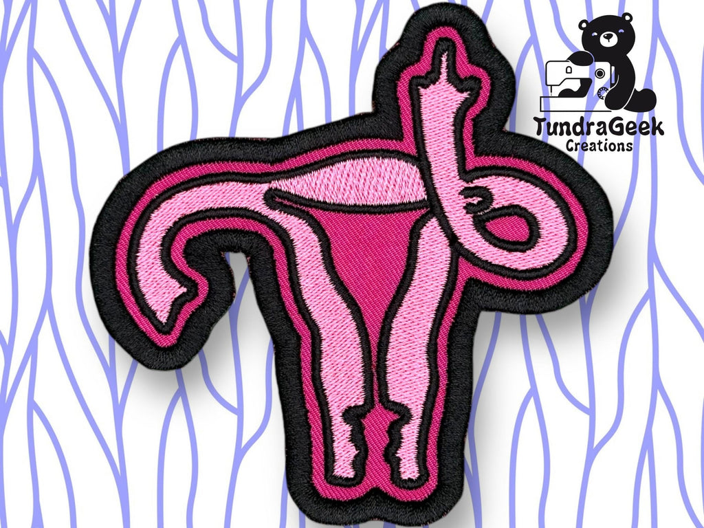 An embroidered pink patch shaped like a uterus with fallopian tubes, outlined in black, displayed on a patterned background. TundraGeek Creations logo is in the top right corner.
