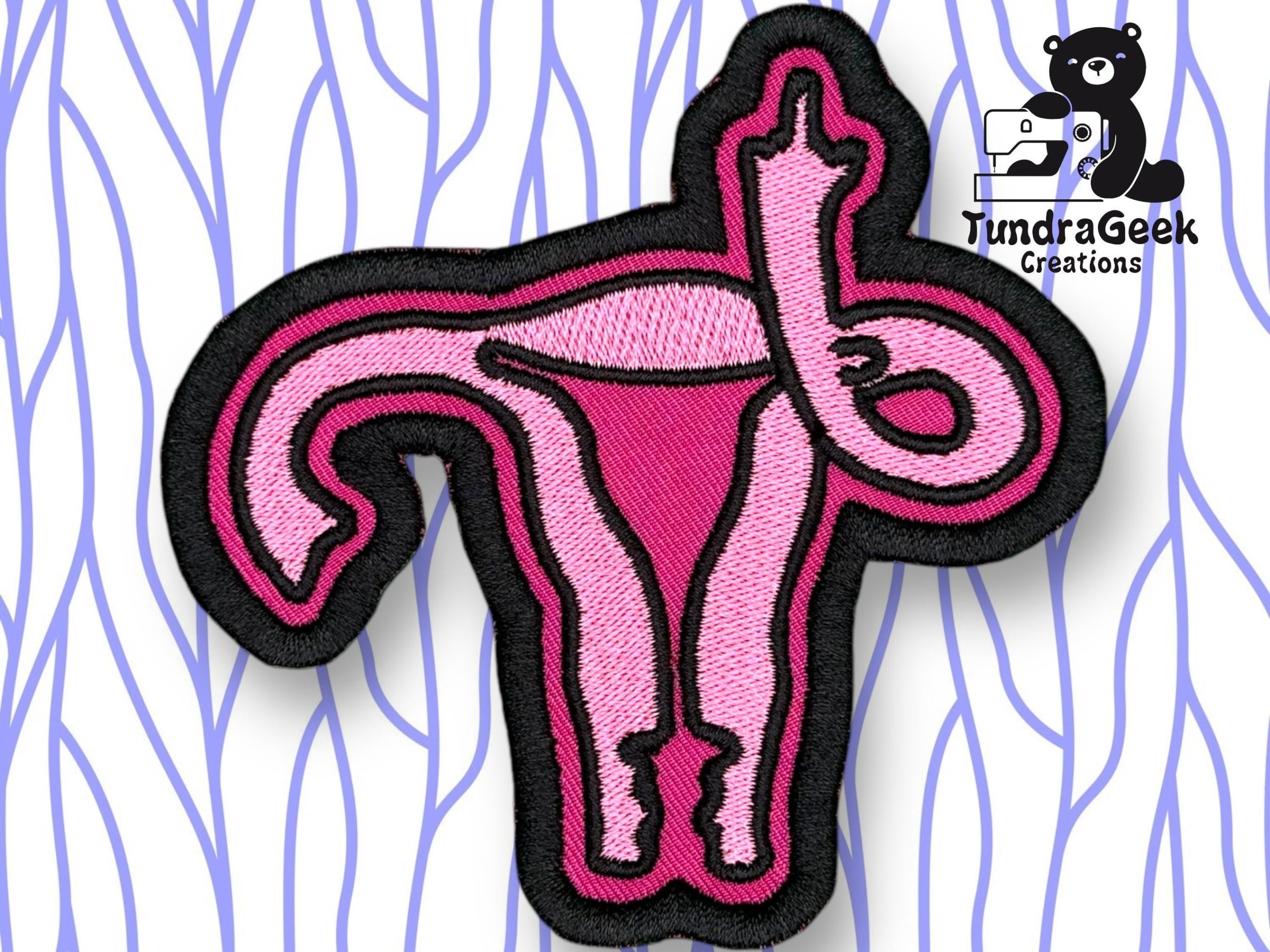 An embroidered pink patch shaped like a uterus with fallopian tubes, outlined in black, displayed on a patterned background. TundraGeek Creations logo is in the top right corner.