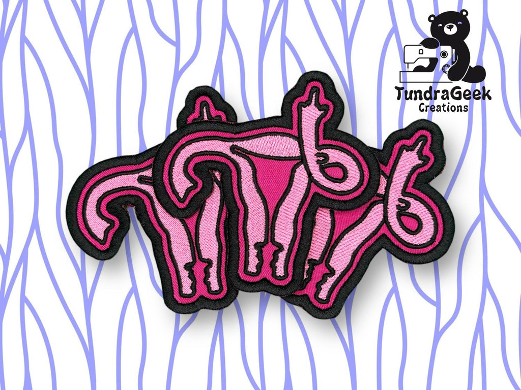 Three embroidered patches depicting pink uteruses overlap on a white background with blue wavy lines, alongside the TundraGeek Creations logo featuring a bear and sewing machine.