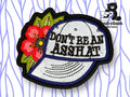 An embroidered patch shows a white baseball cap with the words DONT BE AN ASSHOLE, next to a red flower with green leaves, with a SondraGeek Creations logo in the corner.