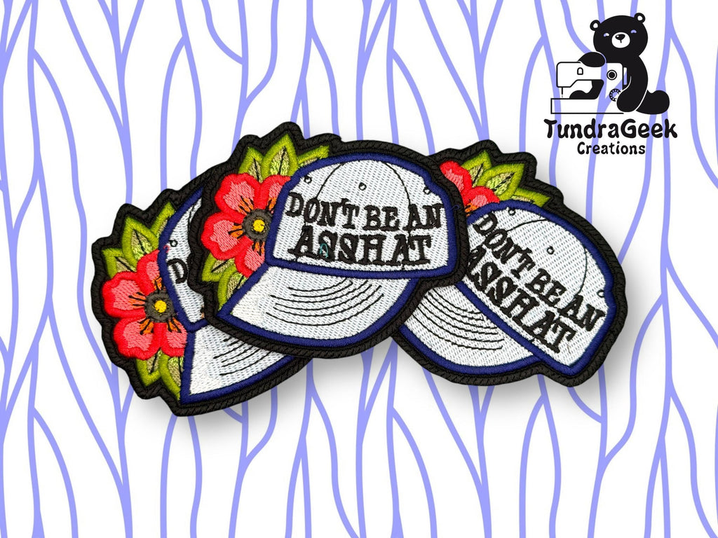 Embroidered patches of hats with red flowers and the phrase DONT BE AN ASSHAT, displayed on a wavy blue and white background with the TundraGeek Creations logo.