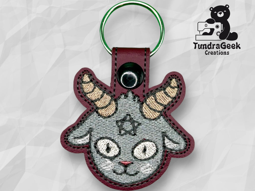 Embroidered keychain featuring a gray goat head with horns, a pentagram on its forehead, and a maroon border. TundraGeek Creations logo appears in the top right corner.
