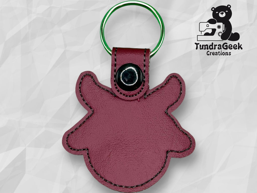 Maroon leather keychain in the shape of an animal head with a snap button, displayed on a geometric background with TundraGeek Creations logo in the top right corner.