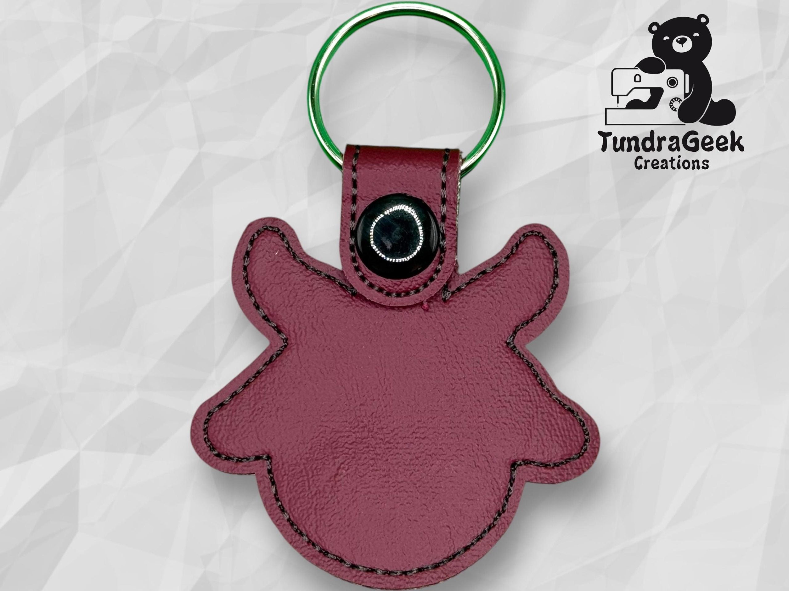 Maroon leather keychain in the shape of an animal head with a snap button, displayed on a geometric background with TundraGeek Creations logo in the top right corner.