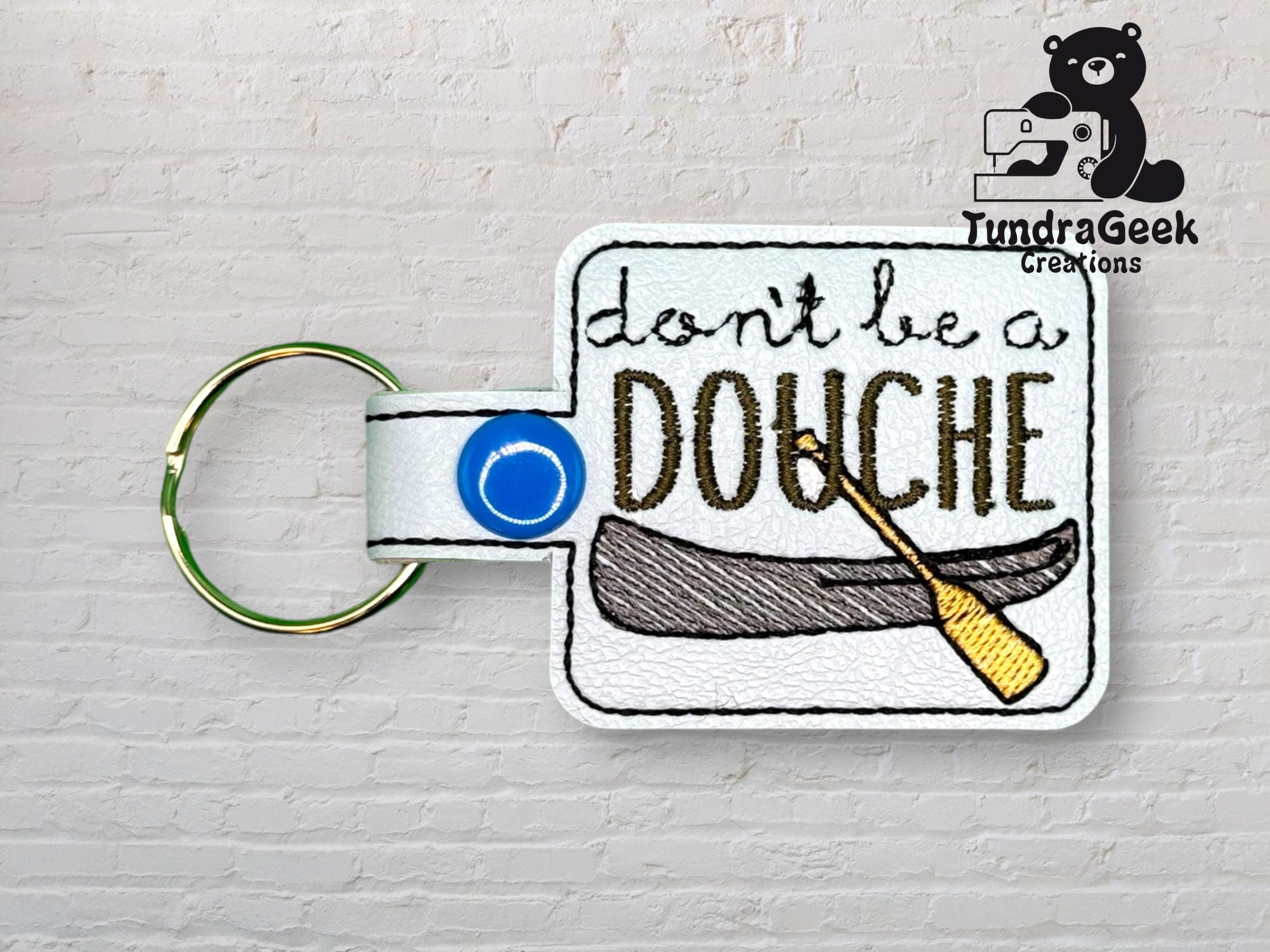 A keychain with embroidered text dont be a DOUCHE above an image of a canoe and a paddle, on a white background with a blue snap button. TundraGeek Creations logo appears in the corner.
