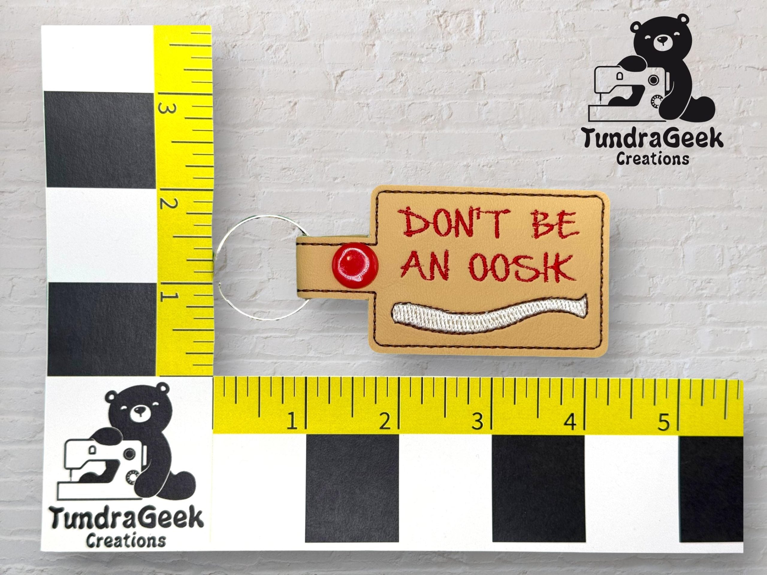 A keychain reads DONT BE AN OOSH in red text, displayed on a measuring grid with TundraGeek Creations logos featuring a bear holding a sewing machine.