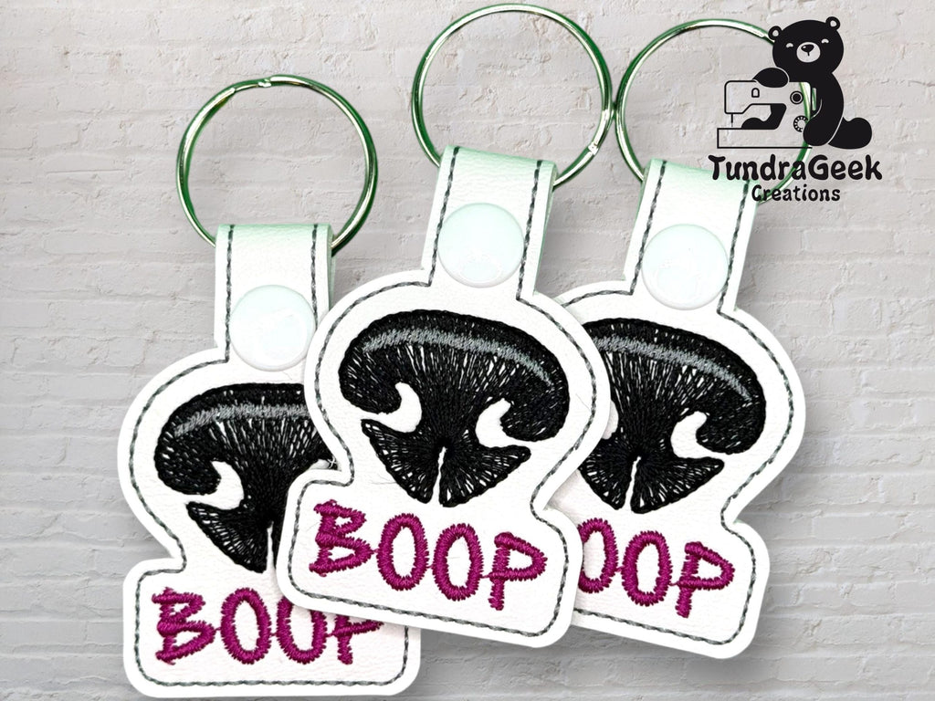 Three keychains with black embroidered dog noses and the word BOOP in purple text, displayed against a light brick background with a TundraGeek Creations logo.