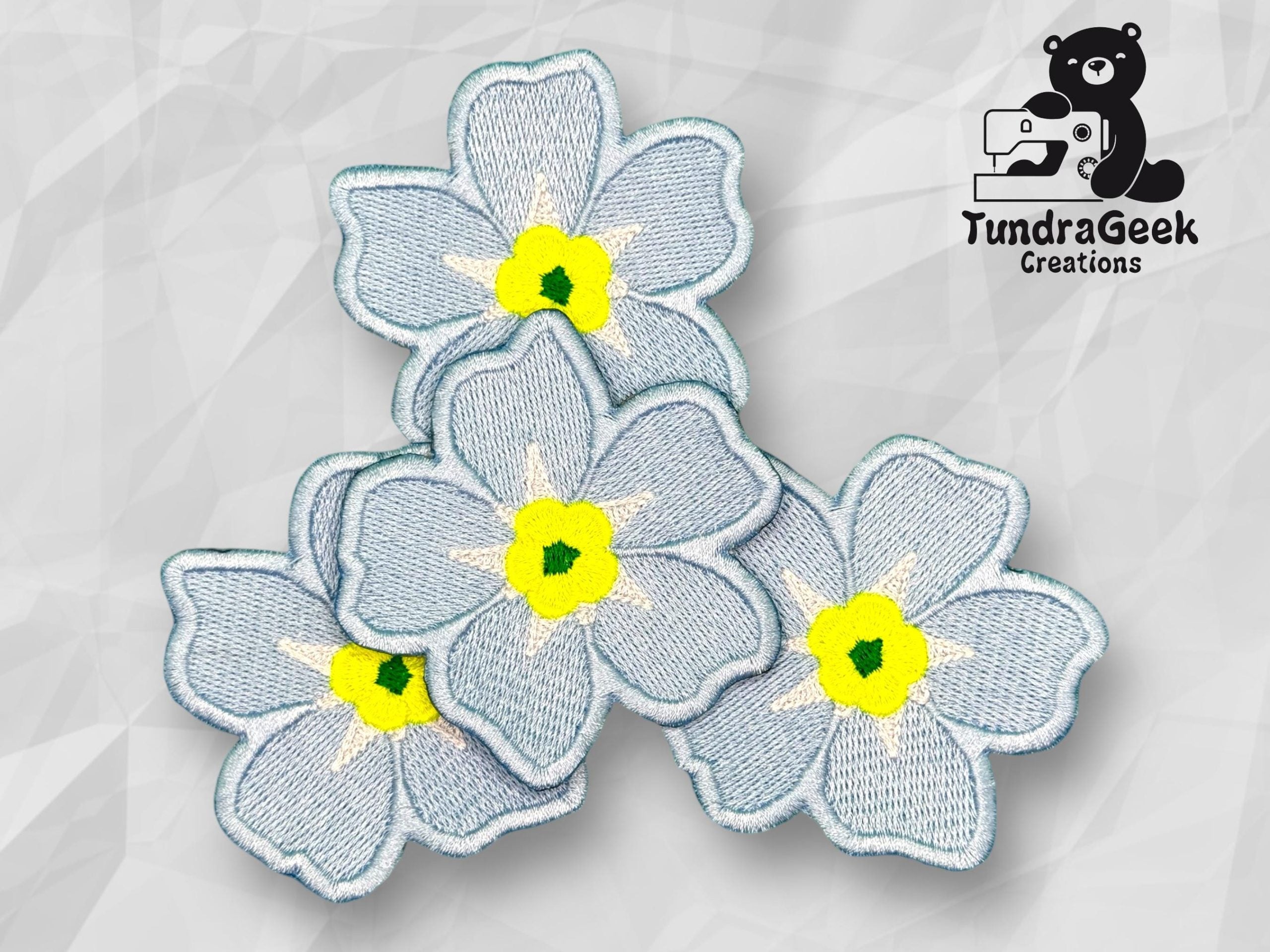 Four light blue embroidered flowers with yellow centers are arranged on a geometric gray background. A logo with a bear and the text TundraGeek Creations is in the top right corner.