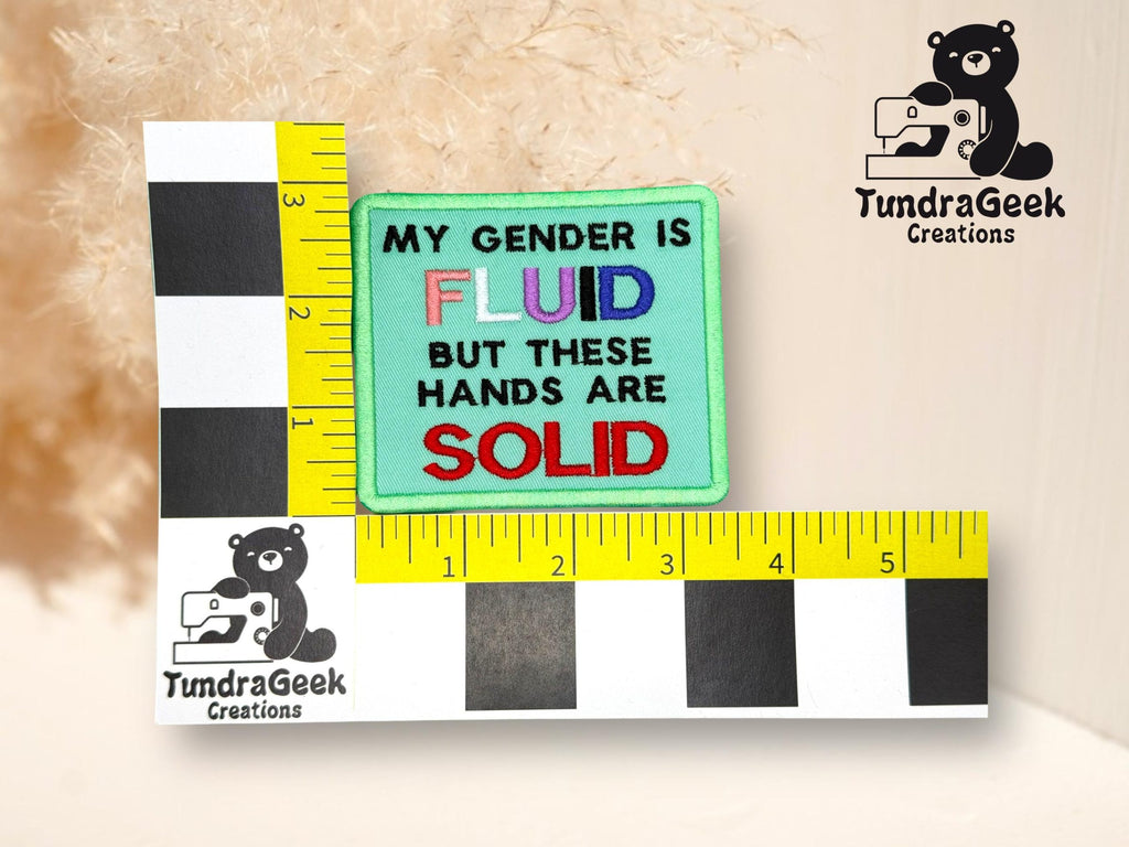 A square patch reads My gender is fluid but these hands are solid with fluid in rainbow colors and solid in red; ruler and TundraGeek Creations logo surround it.
