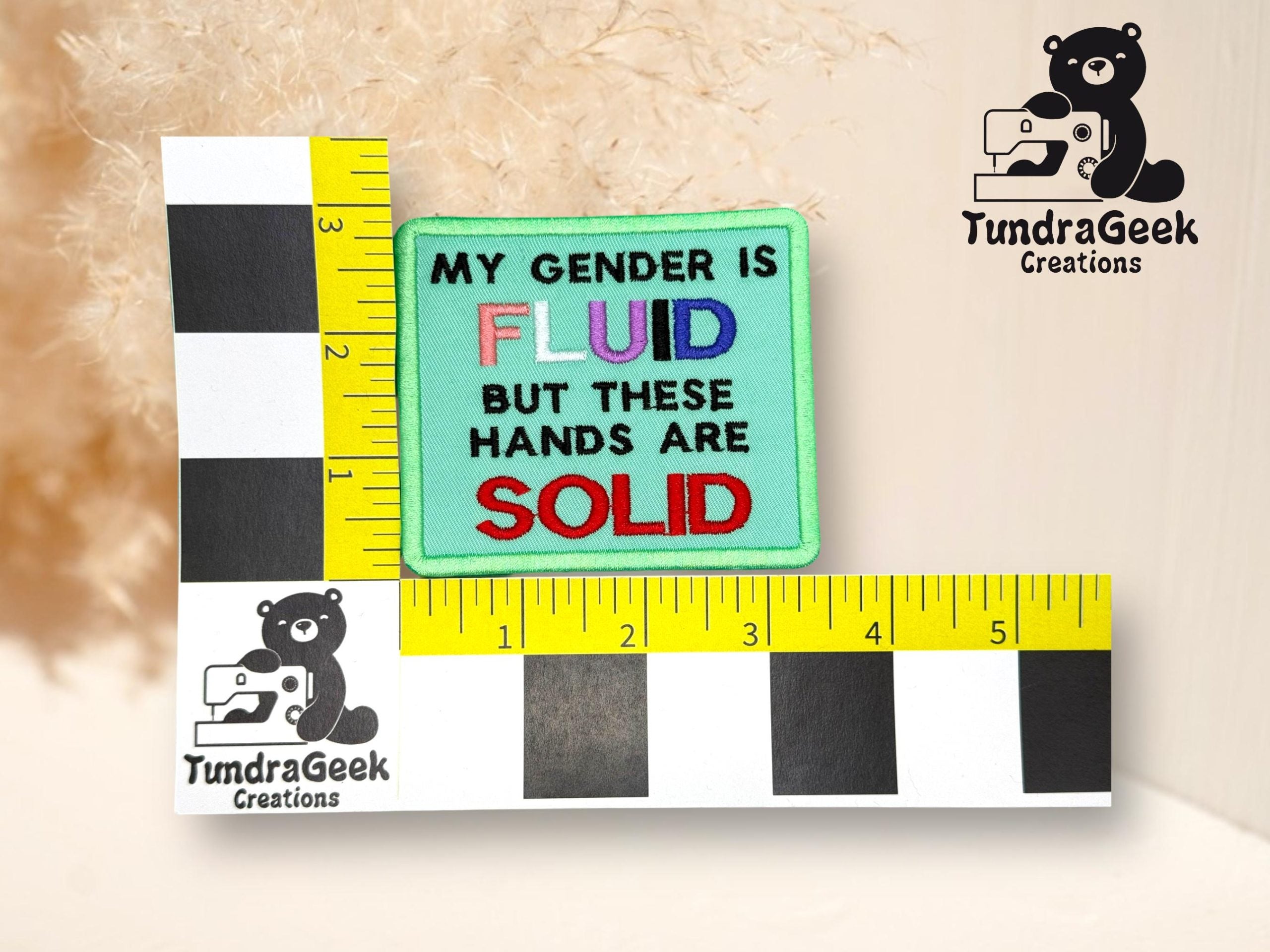 A square patch reads My gender is fluid but these hands are solid with fluid in rainbow colors and solid in red; ruler and TundraGeek Creations logo surround it.