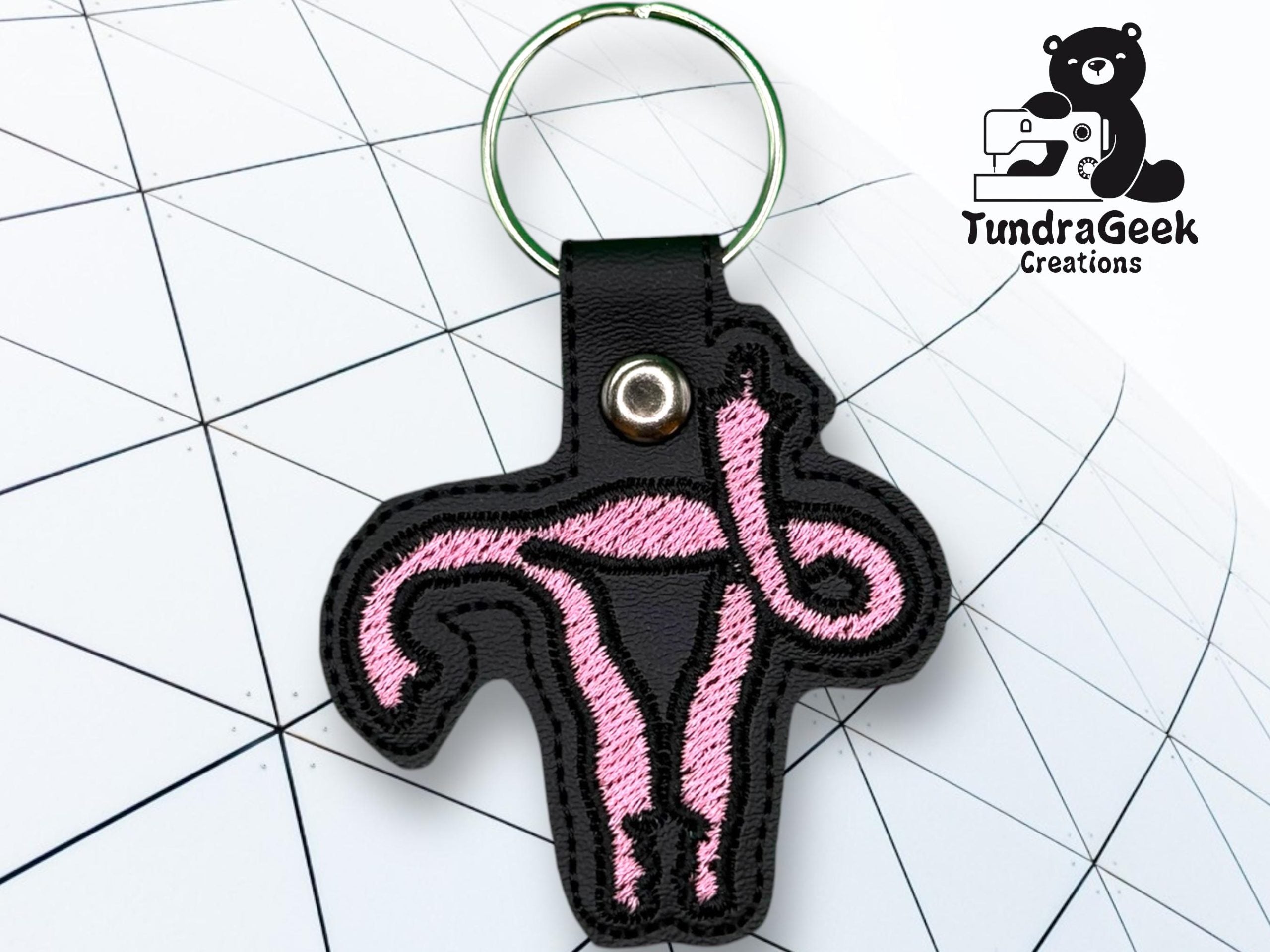 A black keychain with a pink embroidered design of a uterus shape, placed on a grid-patterned background with the TundraGeek Creations logo in the corner.