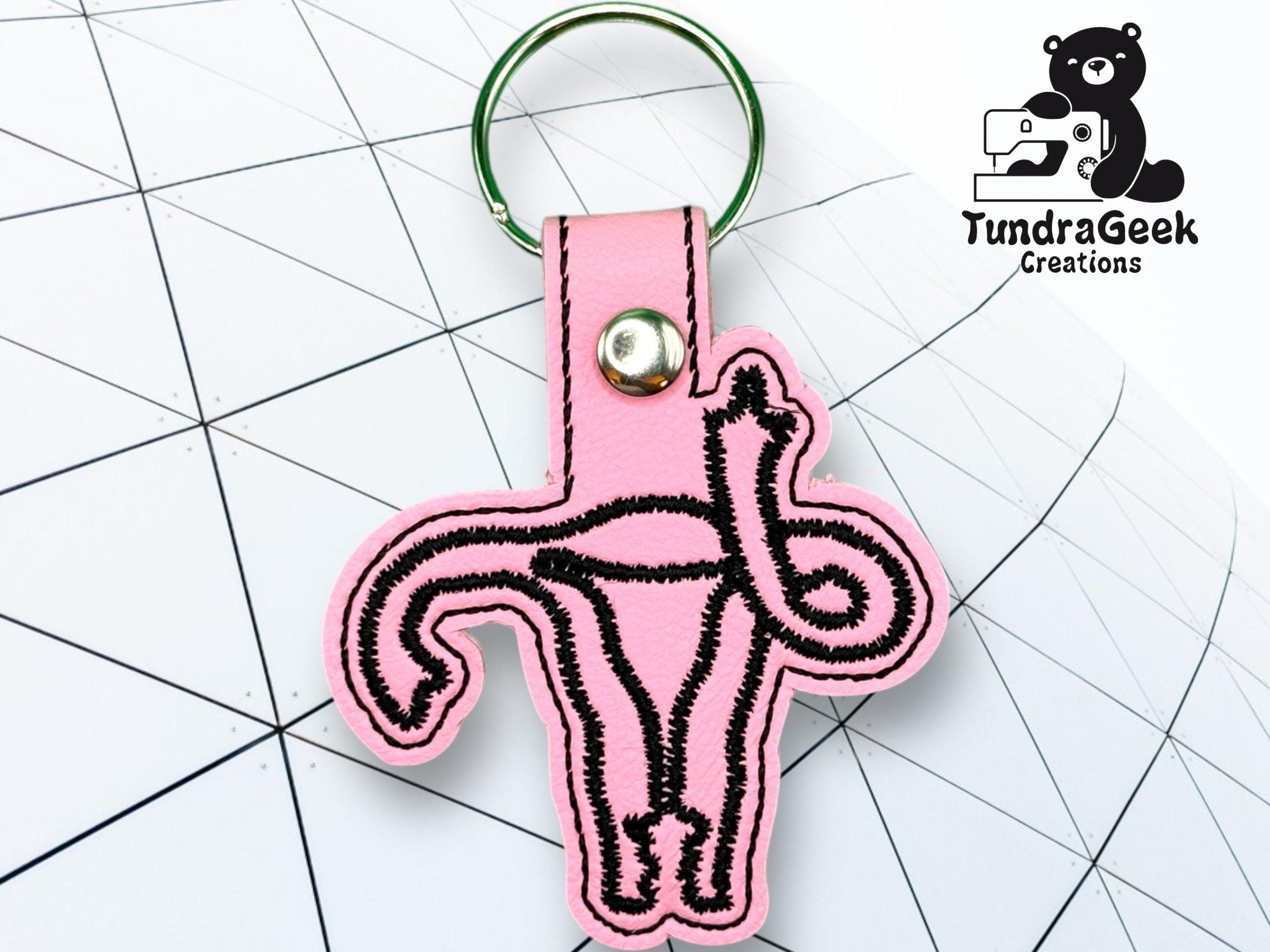 A pink keychain shaped like a uterus with black stitching, placed on a geometric white surface. TundraGeek Creations logo with a bear holding a sewing machine is in the corner.