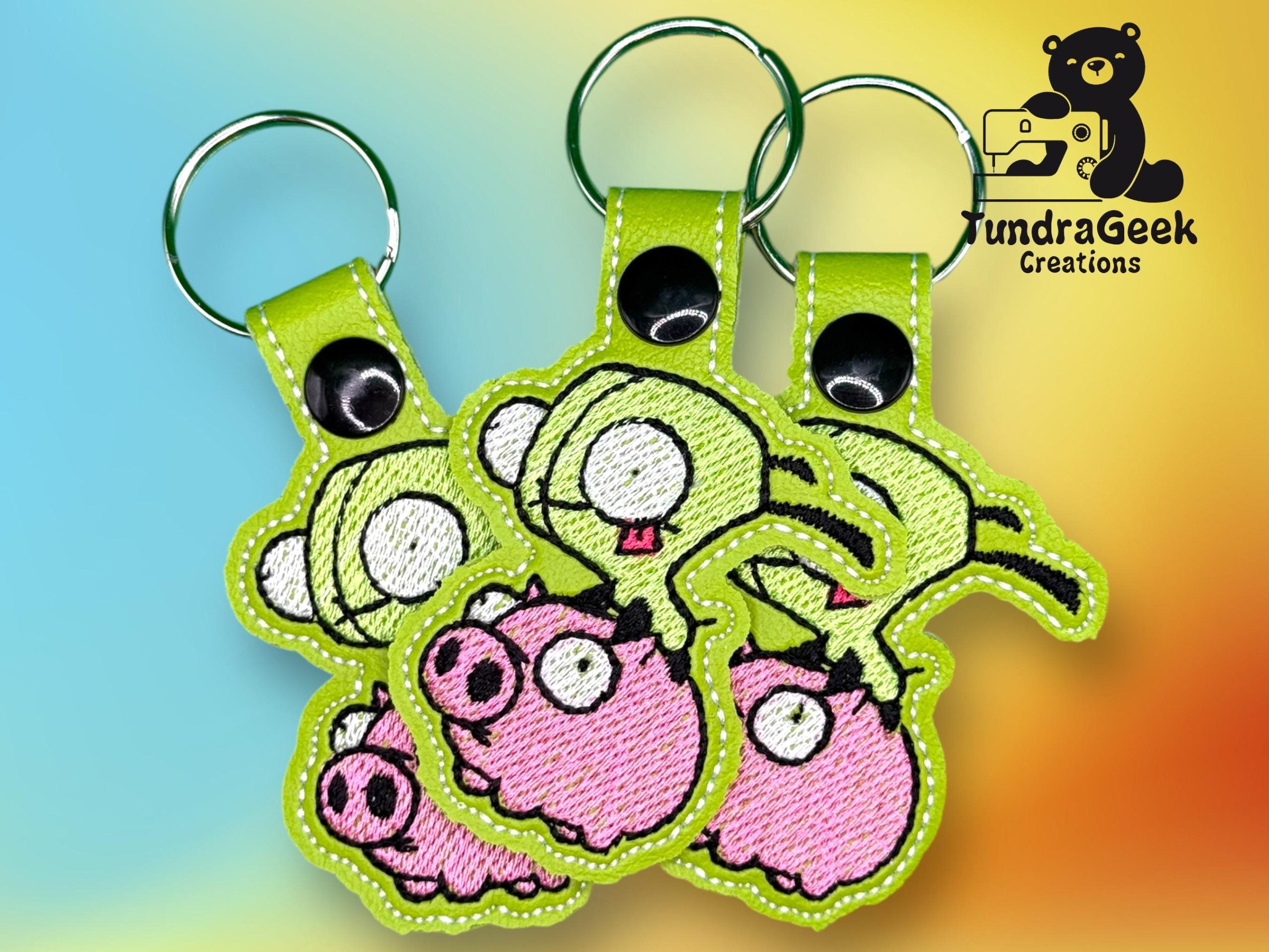 Three embroidered keychains with green and pink pig designs are displayed against a colorful background, featuring a TundraGeek Creations logo in the top right corner.