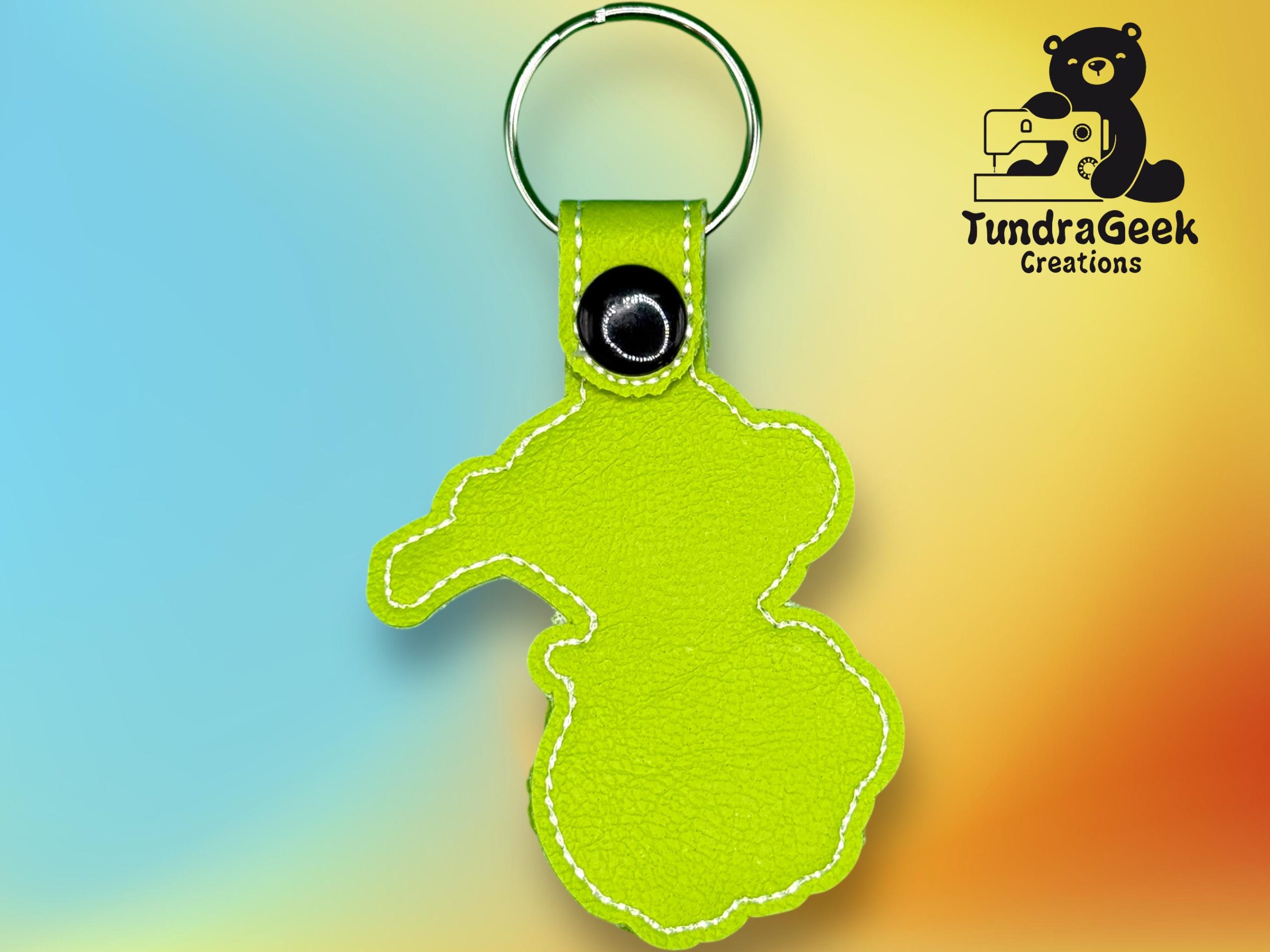 A lime green leather keychain in the shape of a bear with white stitching, metal ring, and a black snap button. Logo for TundraGeek Creations is in the upper right corner.
