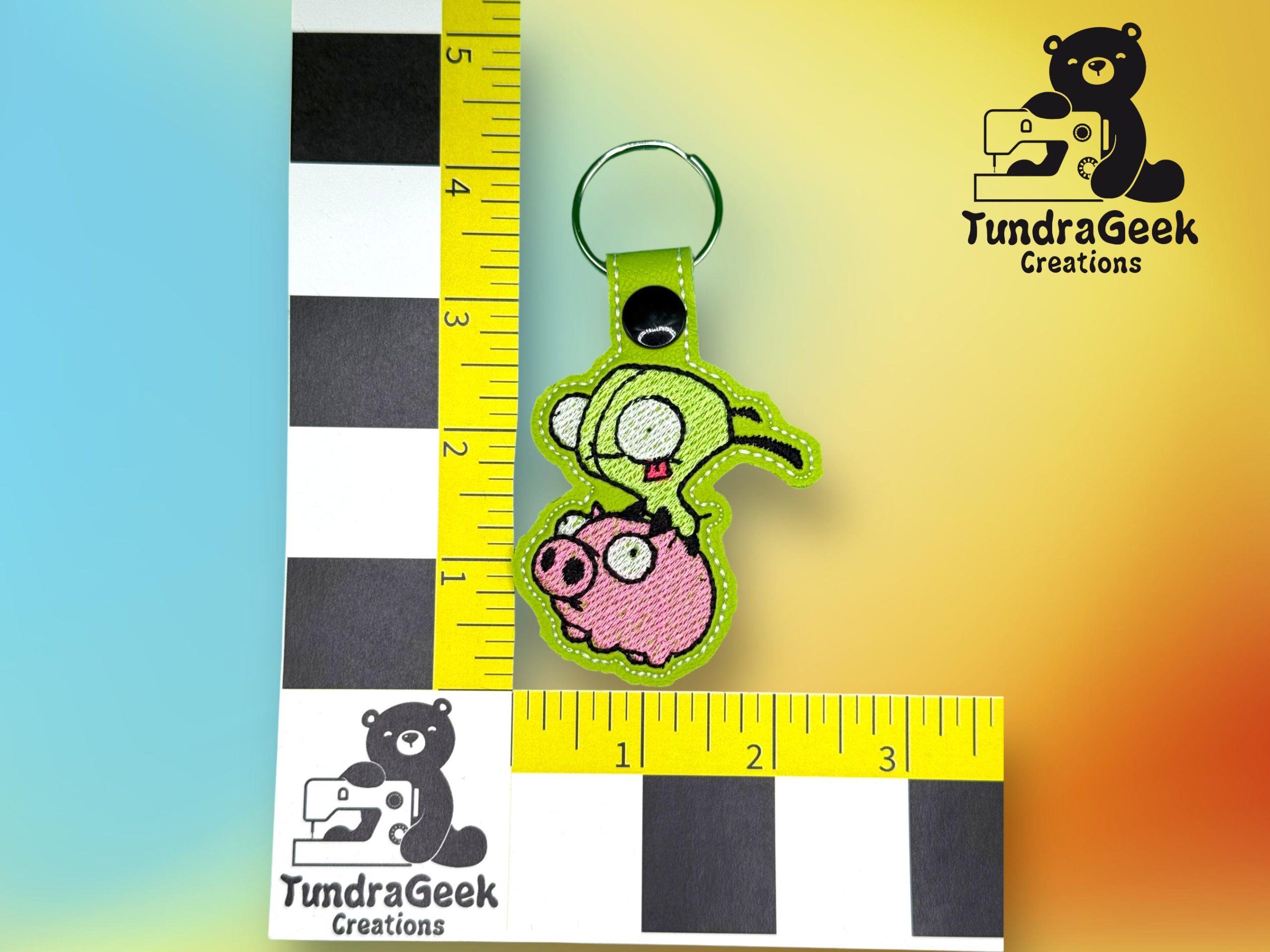 Embroidered keychain with a green alien holding a pink pig, placed between two measuring scales, with the TundraGeek Creations logo in the background.