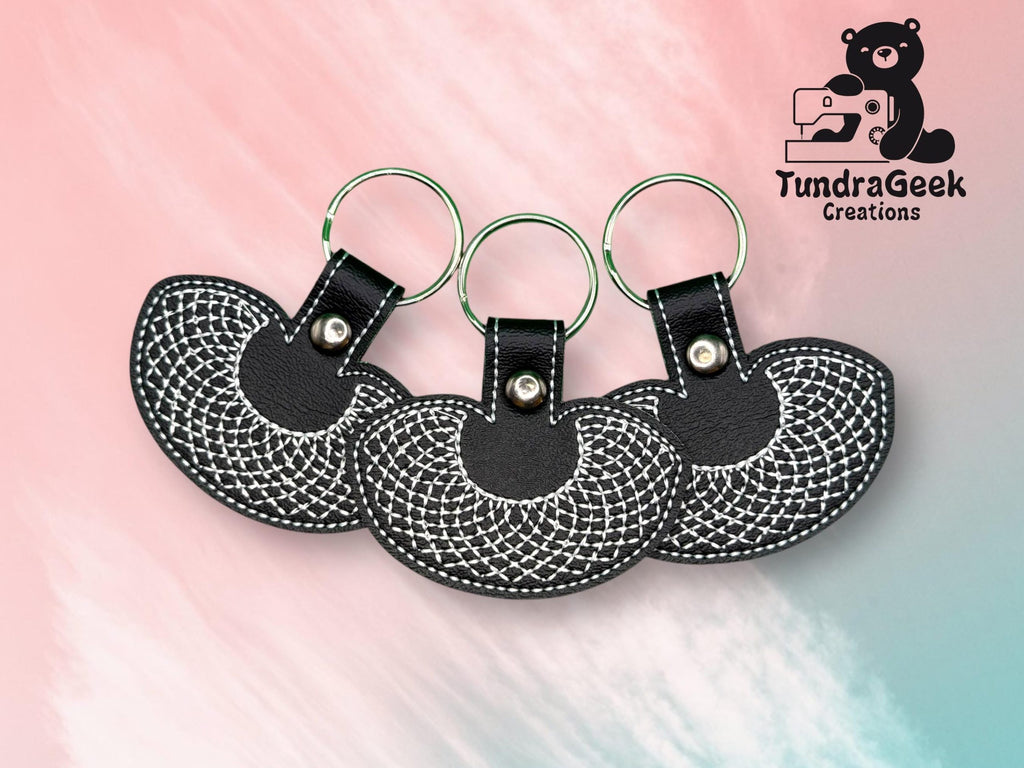 Three black leather keychains with white stitched patterns are arranged in a row on a pink and blue background. The TundraGeek Creations logo is in the top right corner.