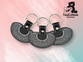 Three black leather keychains with white stitched patterns are arranged in a row on a pink and blue background. The TundraGeek Creations logo is in the top right corner.