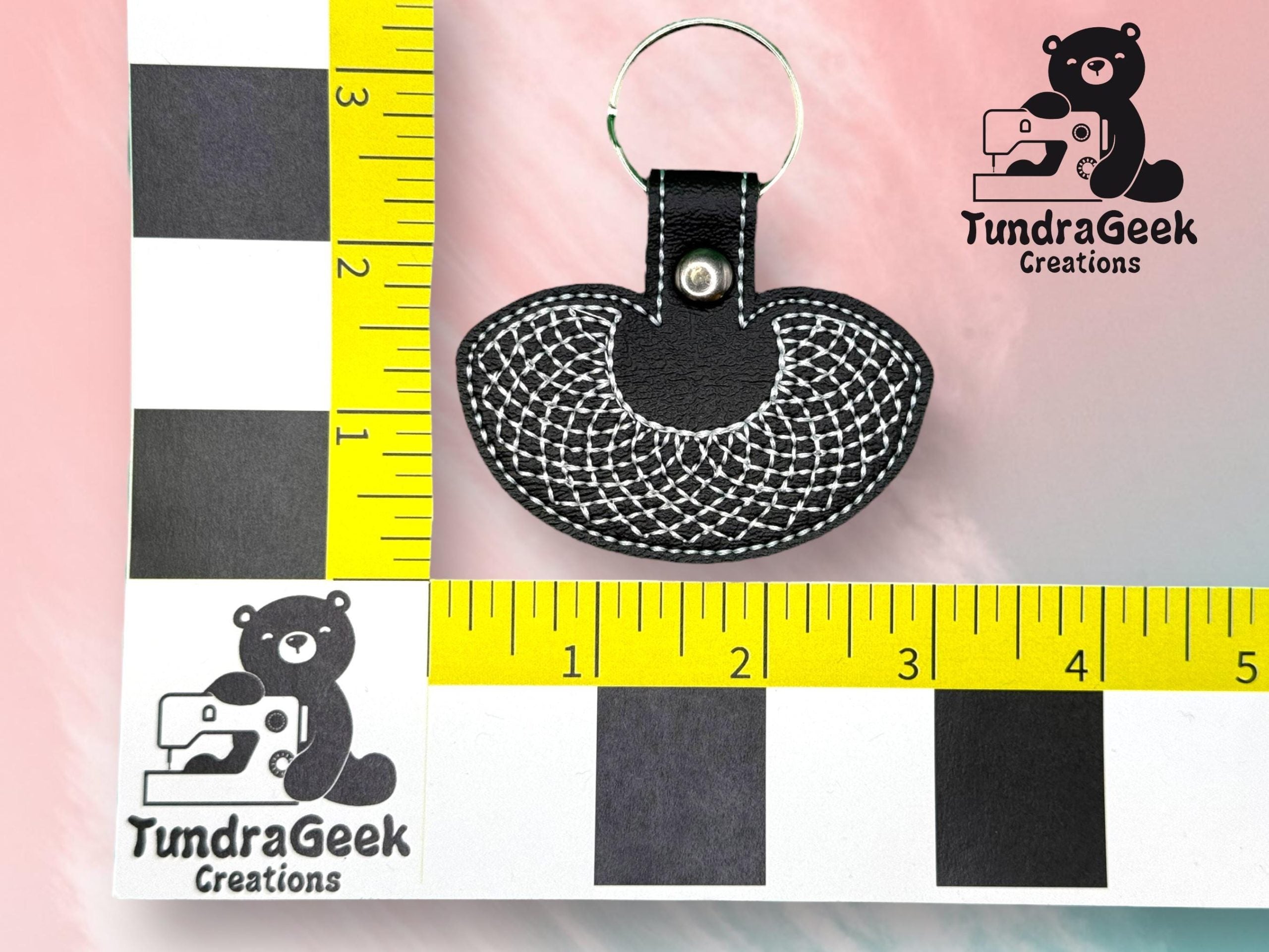 A black and white embroidered keychain is displayed on a measurement grid with the TundraGeek Creations logo featuring a sewing bear in two corners.
