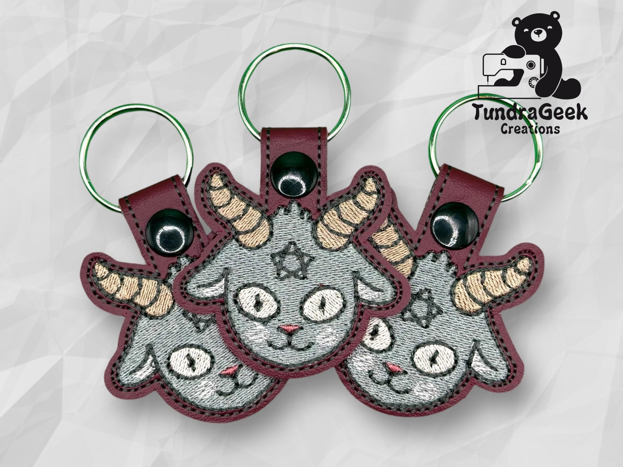 Three embroidered keychains featuring a gray goat head with yellow horns and a pentagram on its forehead, displayed on a geometric background with the TundraGeek Creations logo.