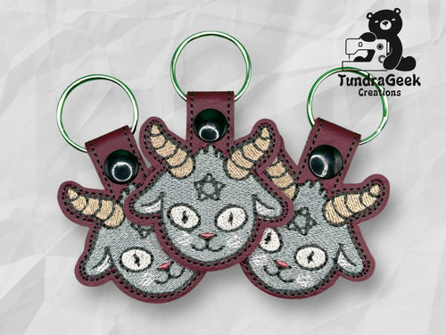 Three embroidered keychains featuring a gray goat head with yellow horns and a pentagram on its forehead, displayed on a geometric background with the TundraGeek Creations logo.