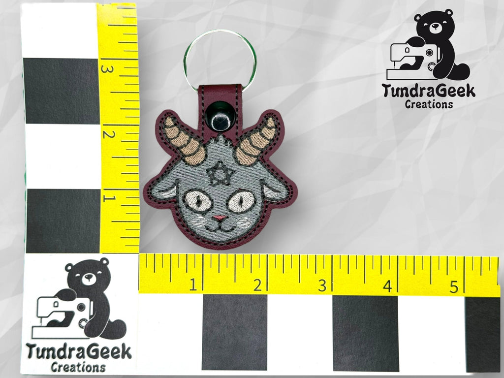 A goat head keychain with embroidered details is placed between two rulers showing measurements, with the TundraGeek Creations logo in the background.