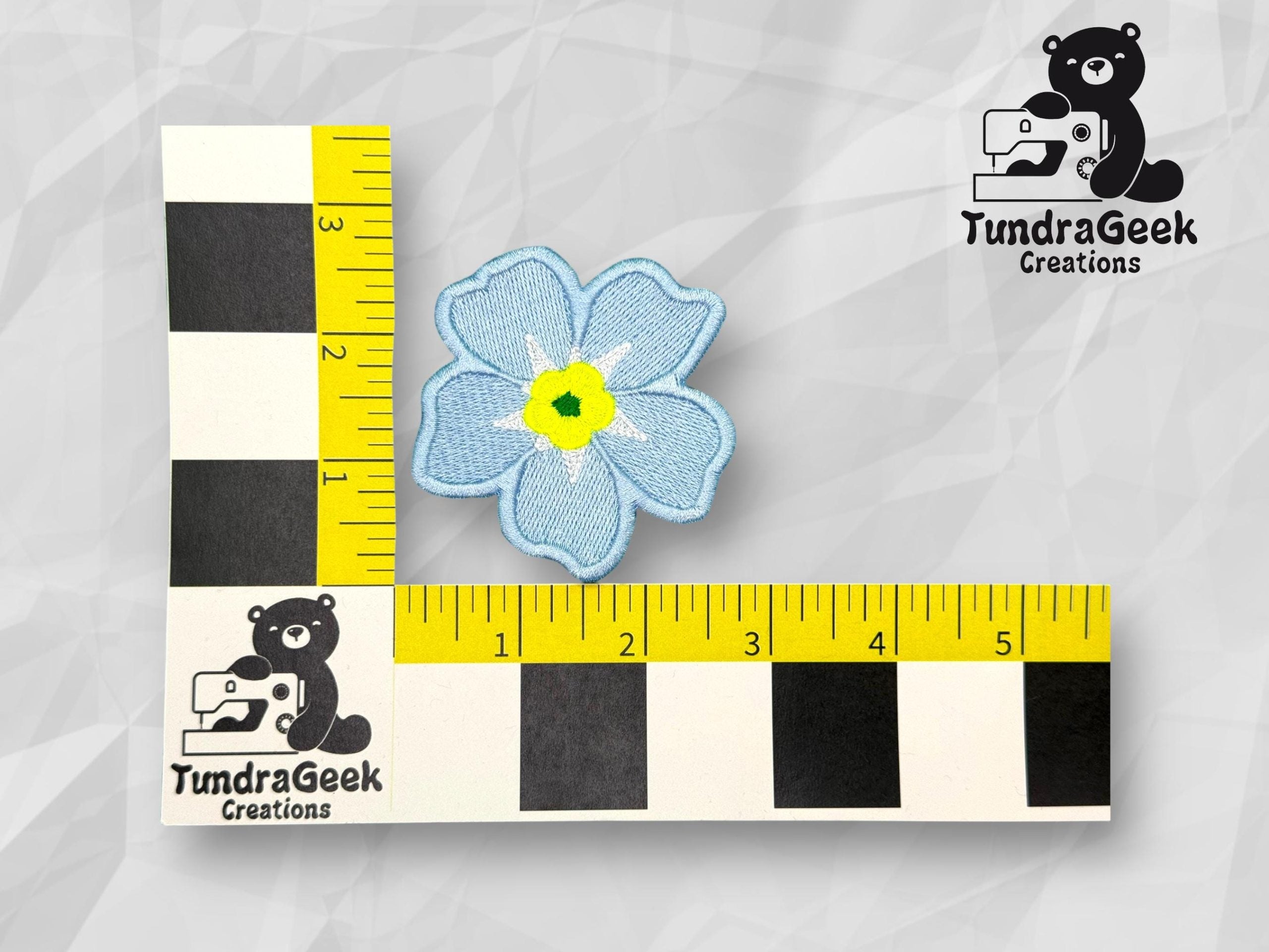 Blue embroidered flower patch with yellow and black center, placed on an L-shaped ruler for scale. TundraGeek Creations logo visible in two corners.