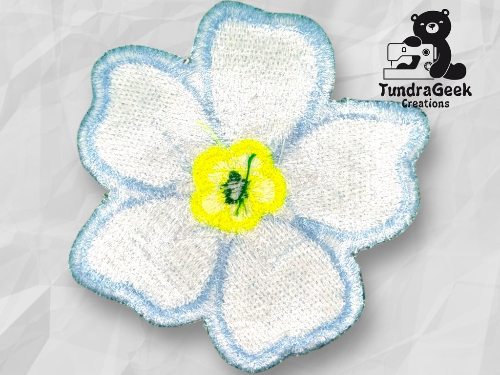 Embroidered white flower patch with blue edges and a yellow center, displayed on a light geometric background with the TundraGeek Creations logo in the top right corner.