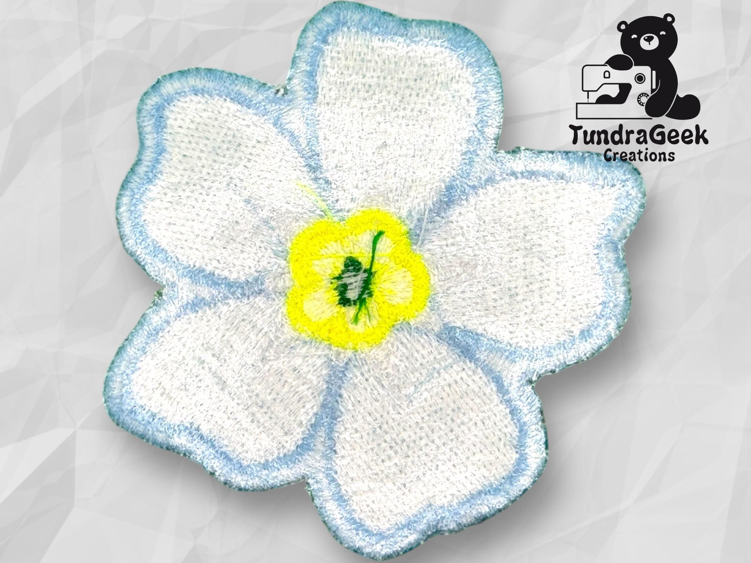 Embroidered white flower patch with blue edges and a yellow center, displayed on a light geometric background with the TundraGeek Creations logo in the top right corner.
