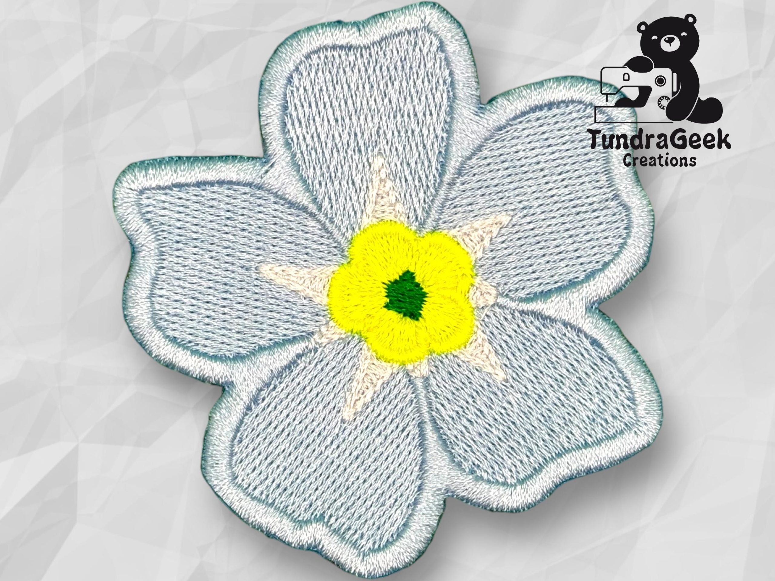Embroidered patch of a blue five-petal flower with a yellow and green center, displayed on a light geometric background with the TundraGeek Creations logo in the top right corner.