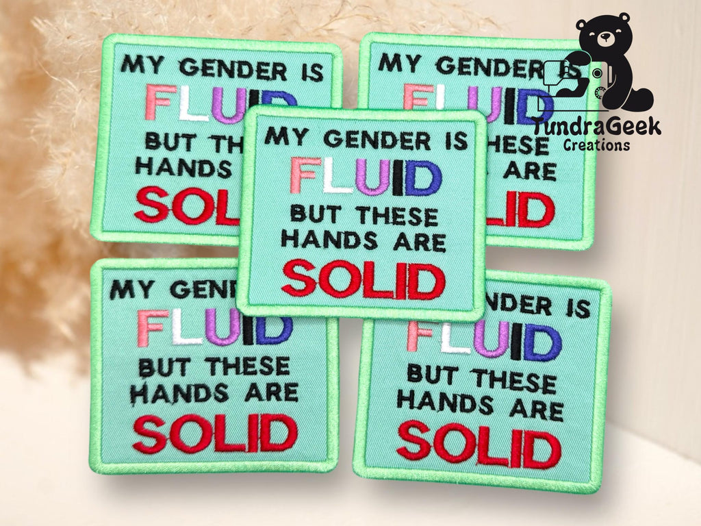 Five embroidered patches with the text: MY GENDER IS FLUID BUT THESE HANDS ARE SOLID in multicolored and bold lettering, displayed on a light surface.