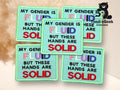 Five embroidered patches with the text: MY GENDER IS FLUID BUT THESE HANDS ARE SOLID in multicolored and bold lettering, displayed on a light surface.