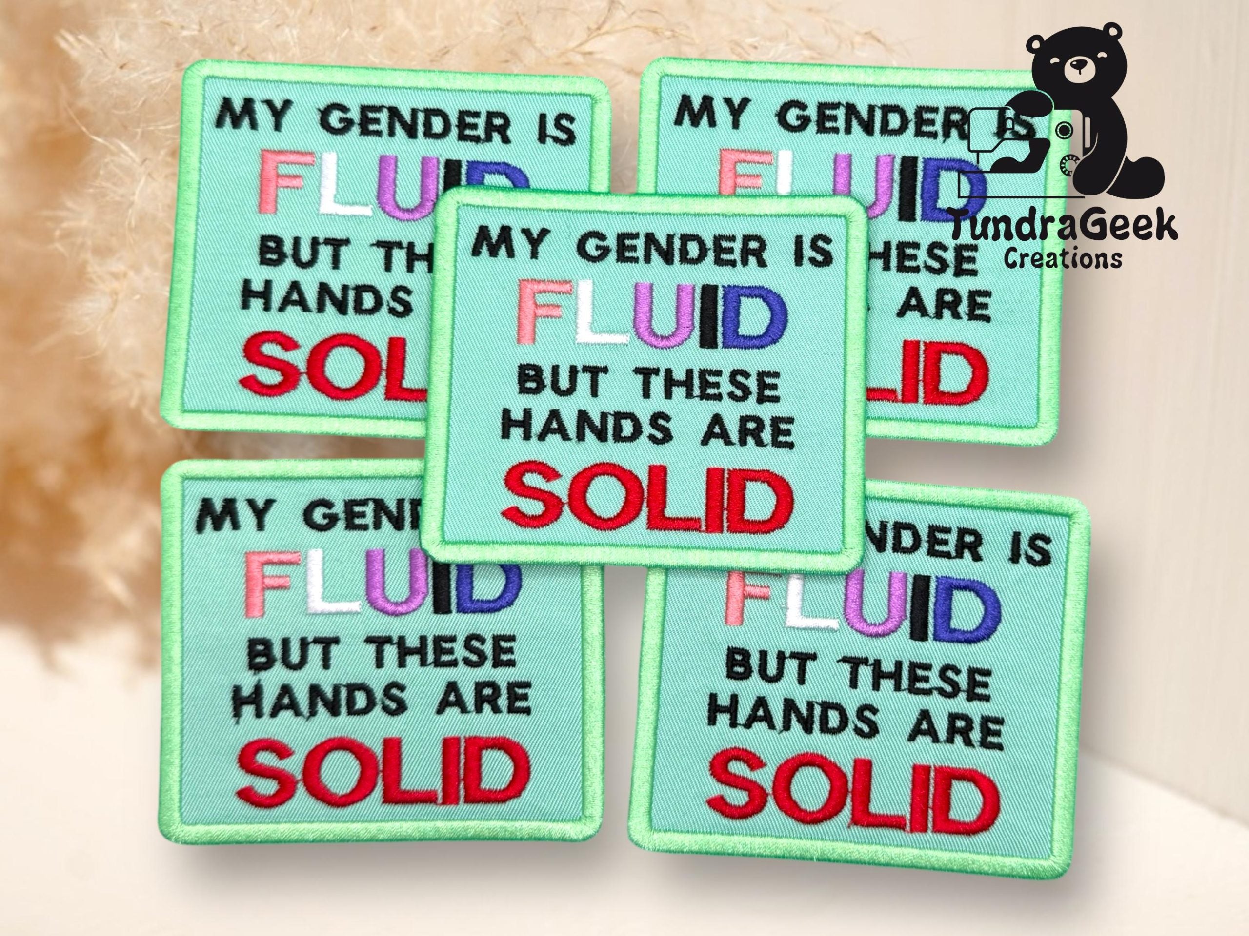 Five fabric patches read: MY GENDER IS FLUID BUT THESE HANDS ARE SOLID with FLUID in multicolored letters and SOLID in red, on a light green background.