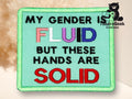 A fabric patch with the text: My gender is FLUID but these hands are SOLID. The word FLUID is in multicolored letters, and SOLID is in red.