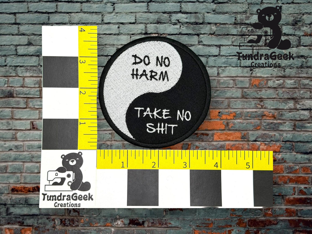 A round black and white patch with the text DO NO HARM TAKE NO SHIT is measured next to a ruler, against a brick wall background.