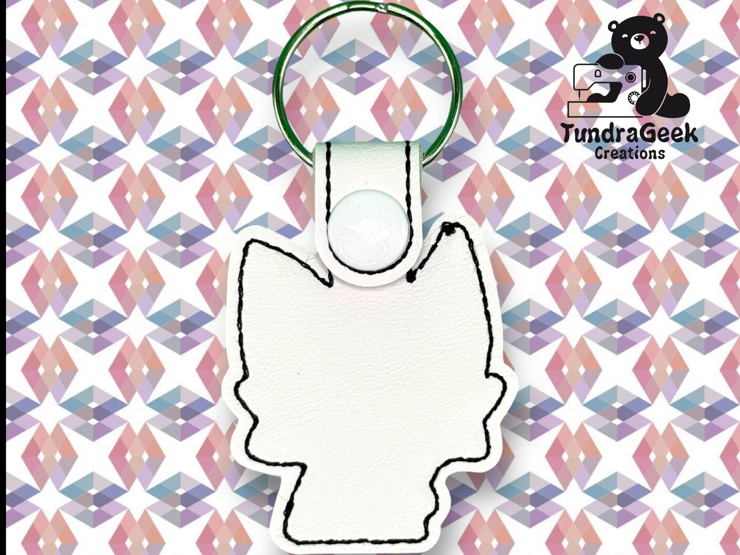 White keychain in the shape of an animal head with black stitching, displayed on a geometric patterned background. TundraGeek Creations logo is in the top right corner.