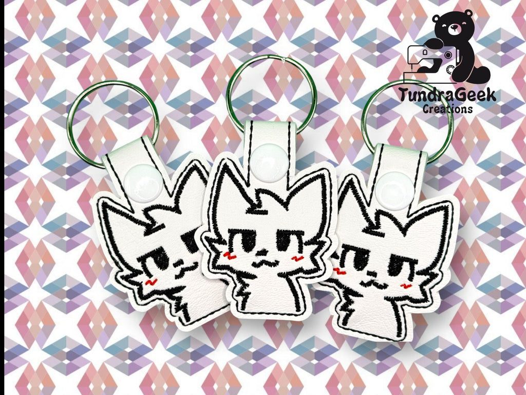 Three identical cat-shaped keychains with embroidered faces and pink cheeks displayed on a geometric patterned background. TundraGeek Creations logo is in the top right corner.