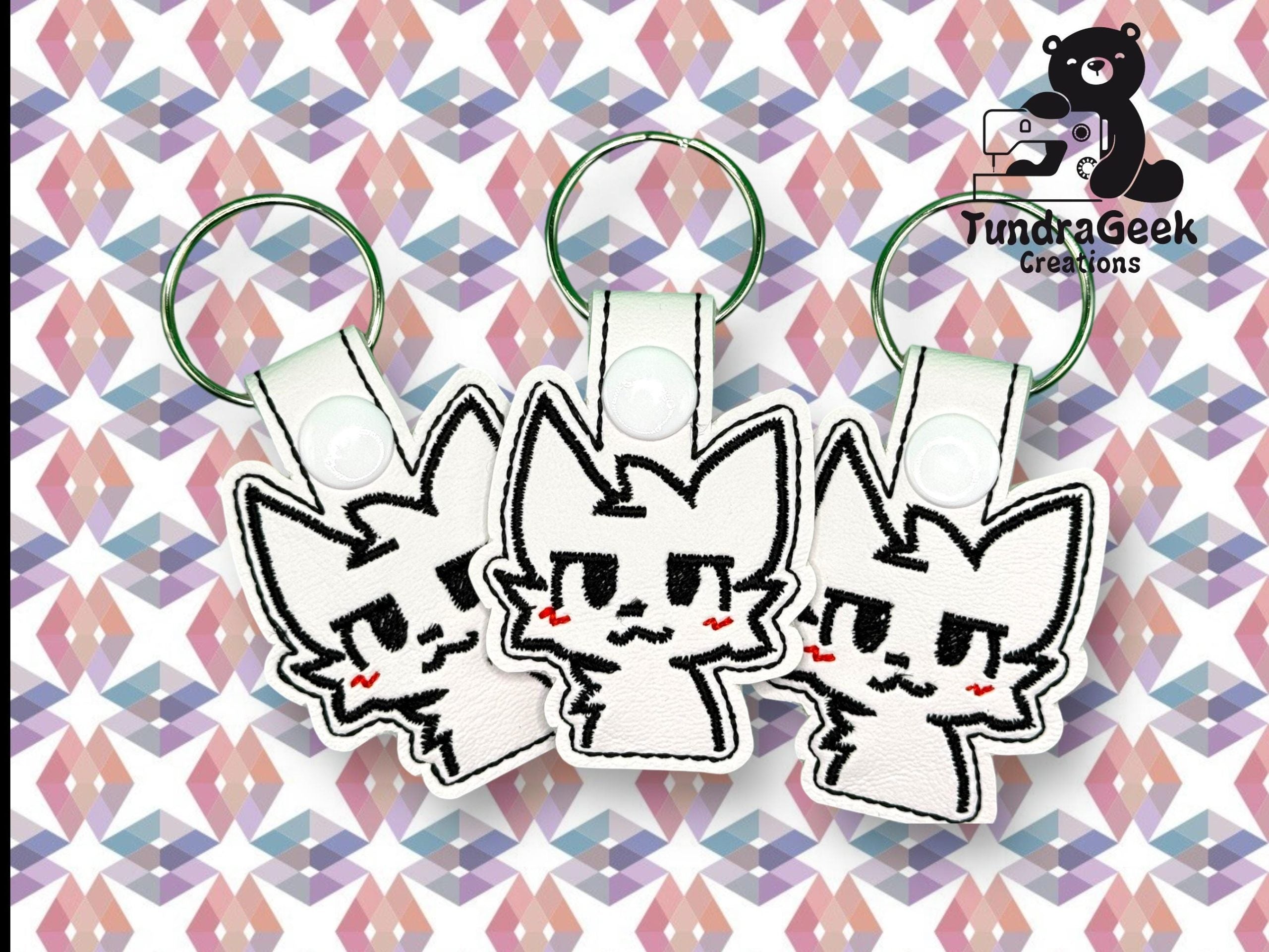 Three identical cat-shaped keychains with embroidered faces and pink cheeks displayed on a geometric patterned background. TundraGeek Creations logo is in the top right corner.