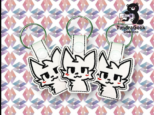 Three identical cat-shaped keychains with embroidered faces and pink cheeks displayed on a geometric patterned background. TundraGeek Creations logo is in the top right corner.