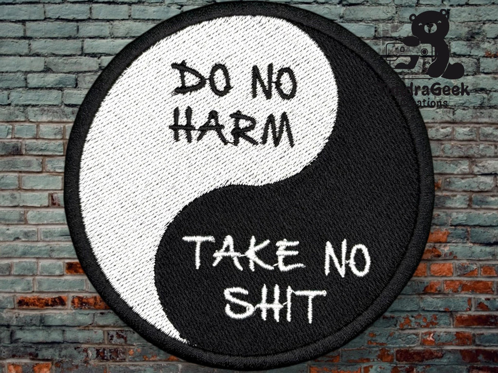 A round yin-yang patch with the text “DO NO HARM” on the white half and “TAKE NO SHIT” on the black half, set against a brick wall background.