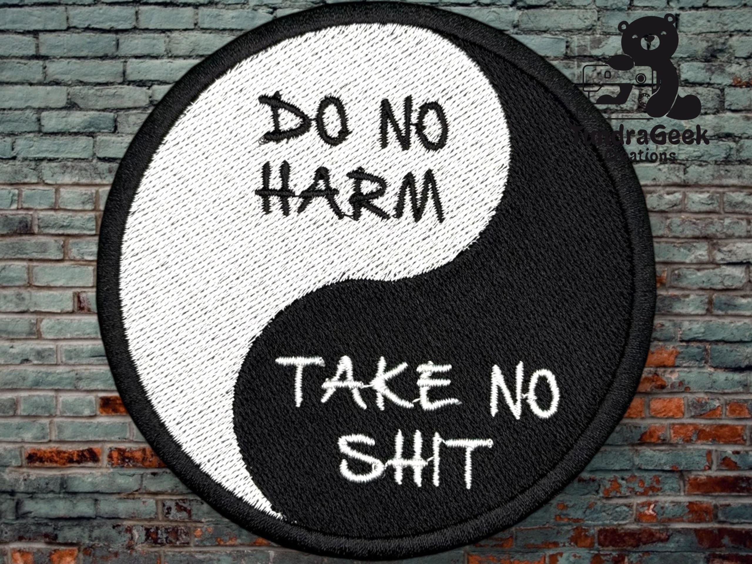 A round yin-yang patch with the text “DO NO HARM” on the white half and “TAKE NO SHIT” on the black half, set against a brick wall background.