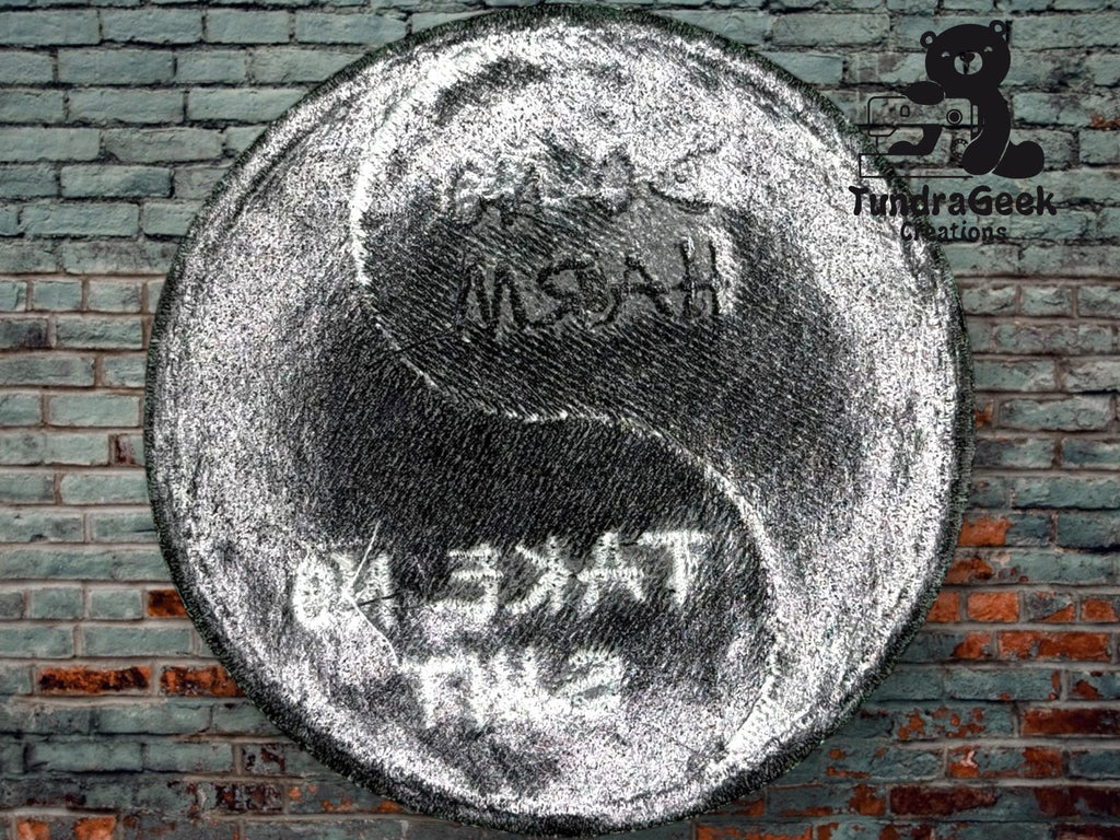 A metallic yin-yang symbol with engraved text is displayed against a brick wall background. The TundraGeek Creations logo appears in the upper right corner.