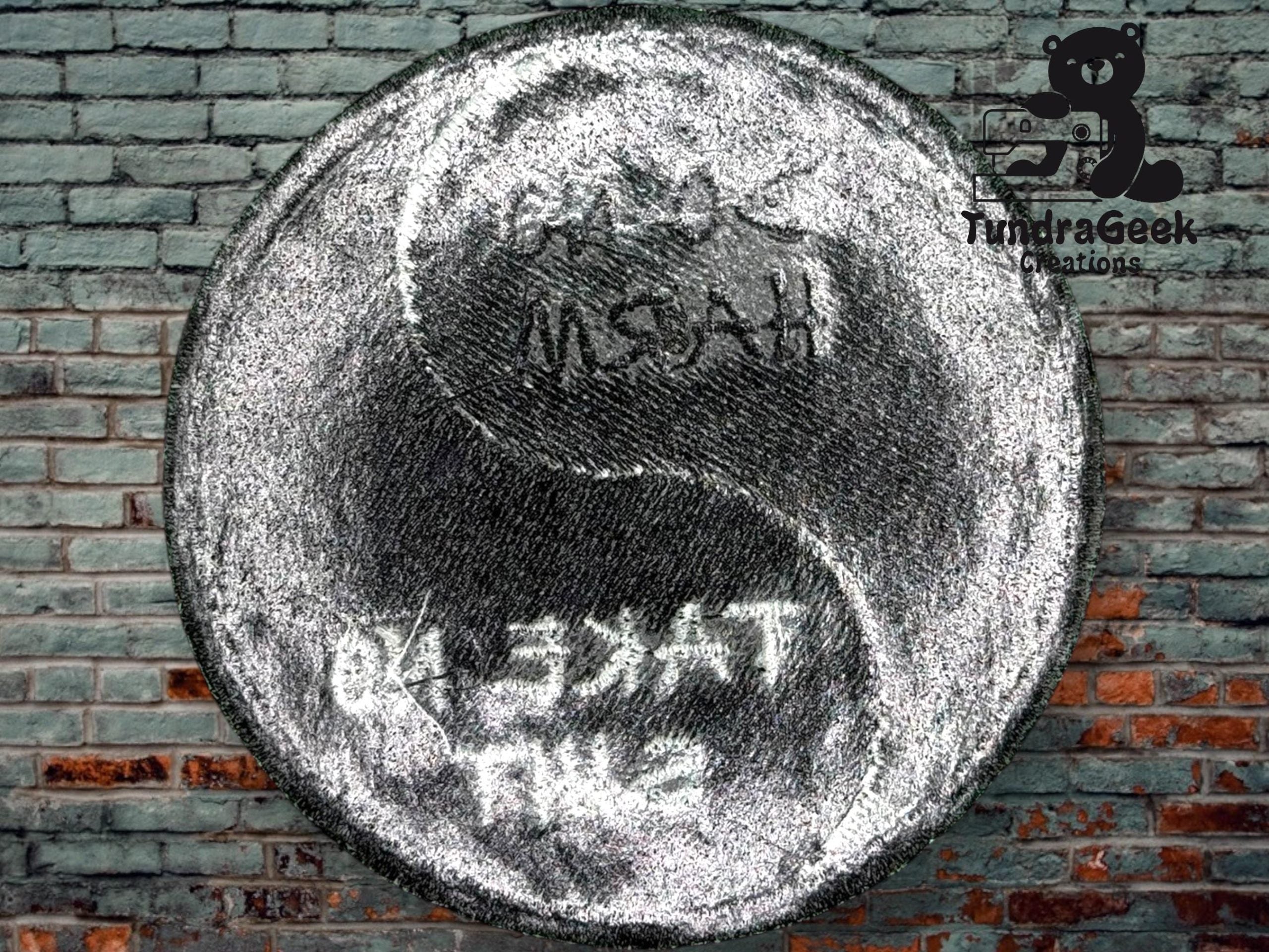 A metallic yin-yang symbol with engraved text is displayed against a brick wall background. The TundraGeek Creations logo appears in the upper right corner.