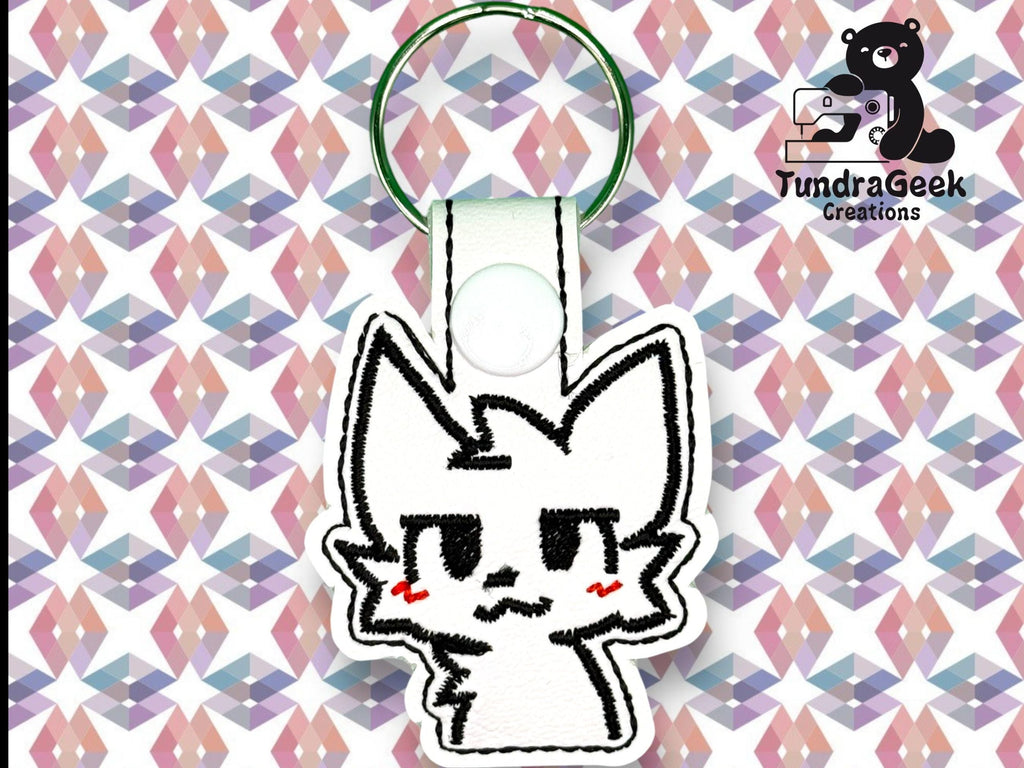 A white keychain with a cartoon cat face and red cheeks is displayed on a geometric pastel background. Logo in the corner reads TundraGeek Creations with a bear and sewing machine icon.