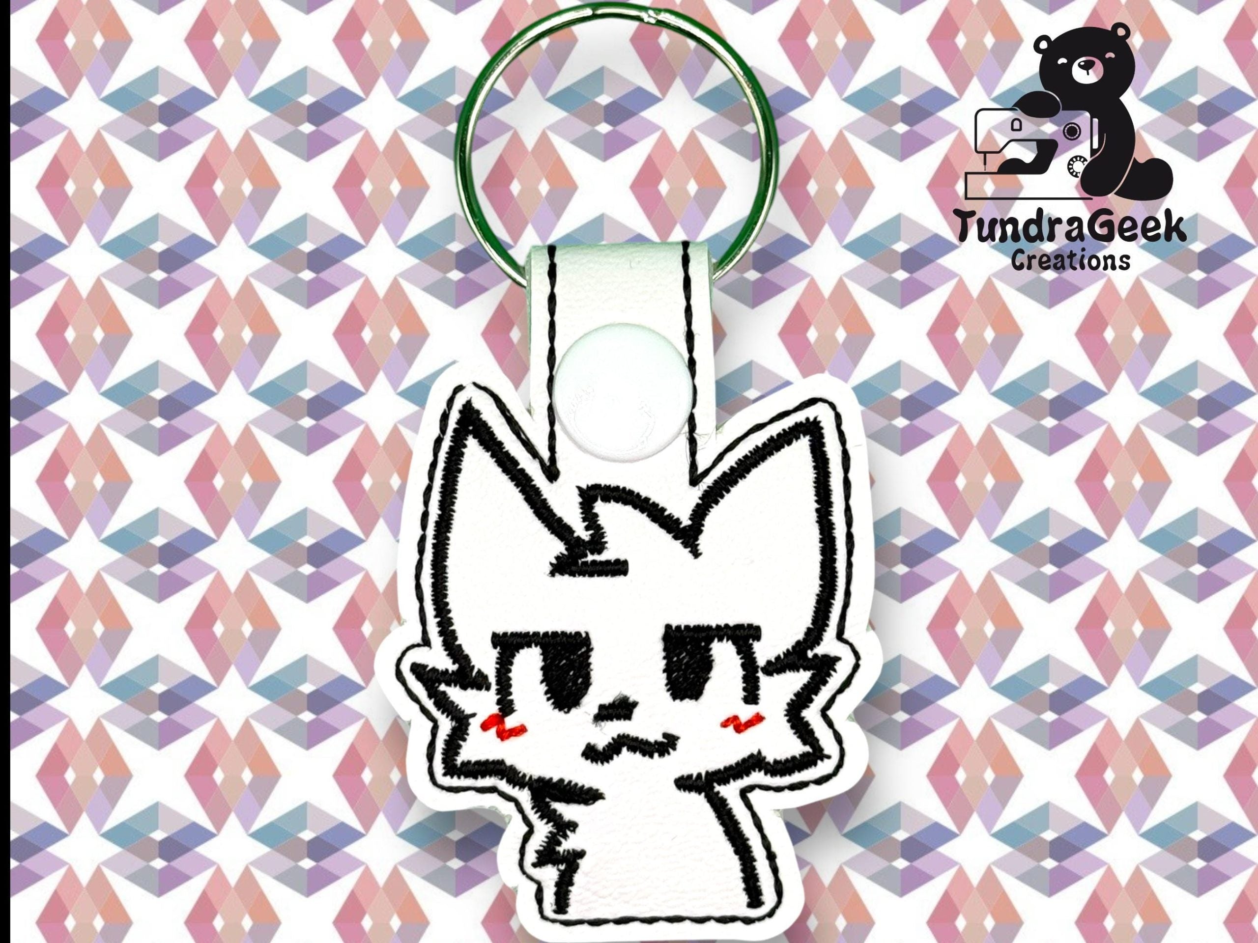 A white keychain with a cartoon cat face and red cheeks is displayed on a geometric pastel background. Logo in the corner reads TundraGeek Creations with a bear and sewing machine icon.