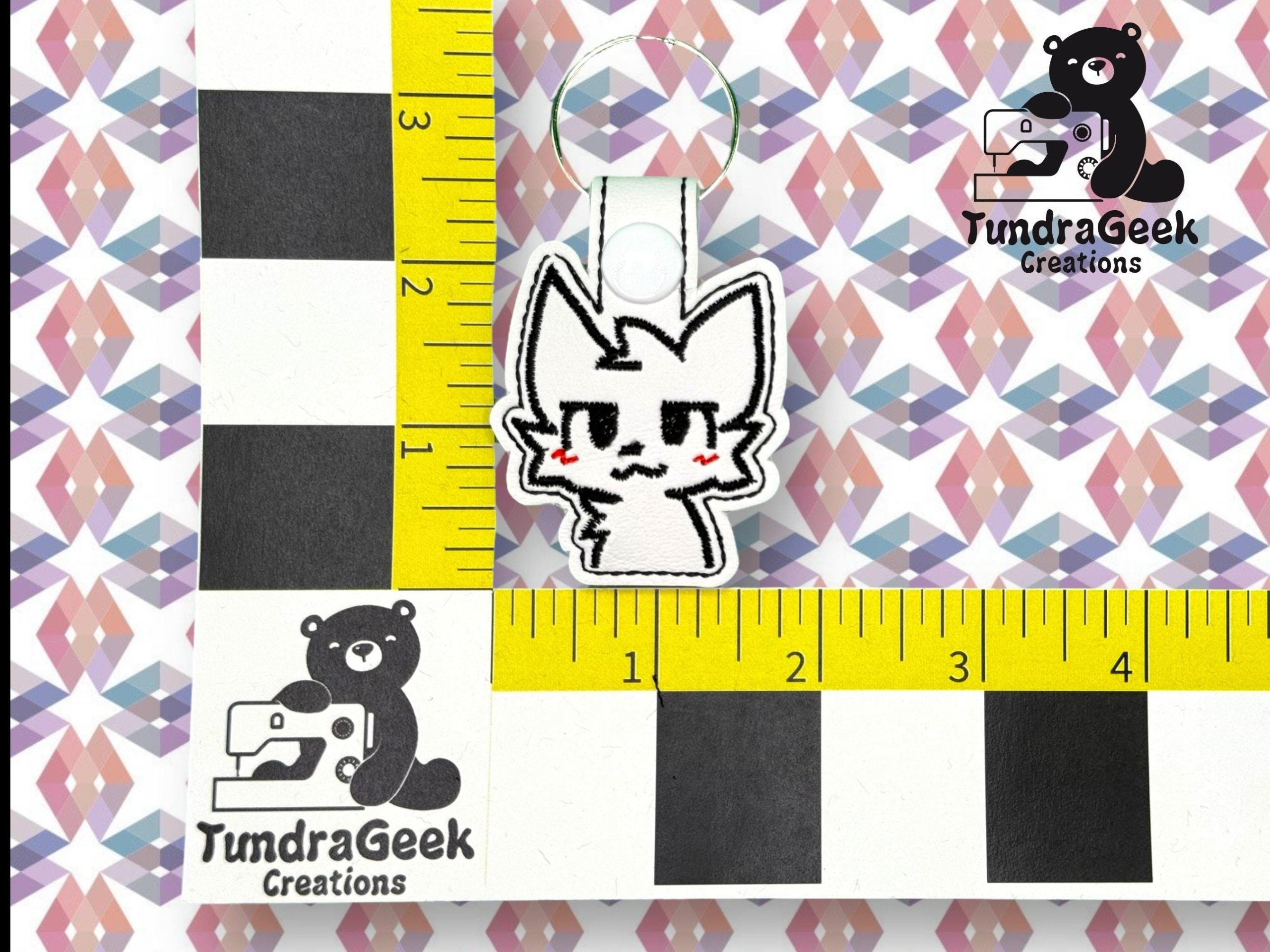 A small keychain with a cartoon animal face stitched on white fabric is placed on a measured grid with TundraGeek Creations bear logos.