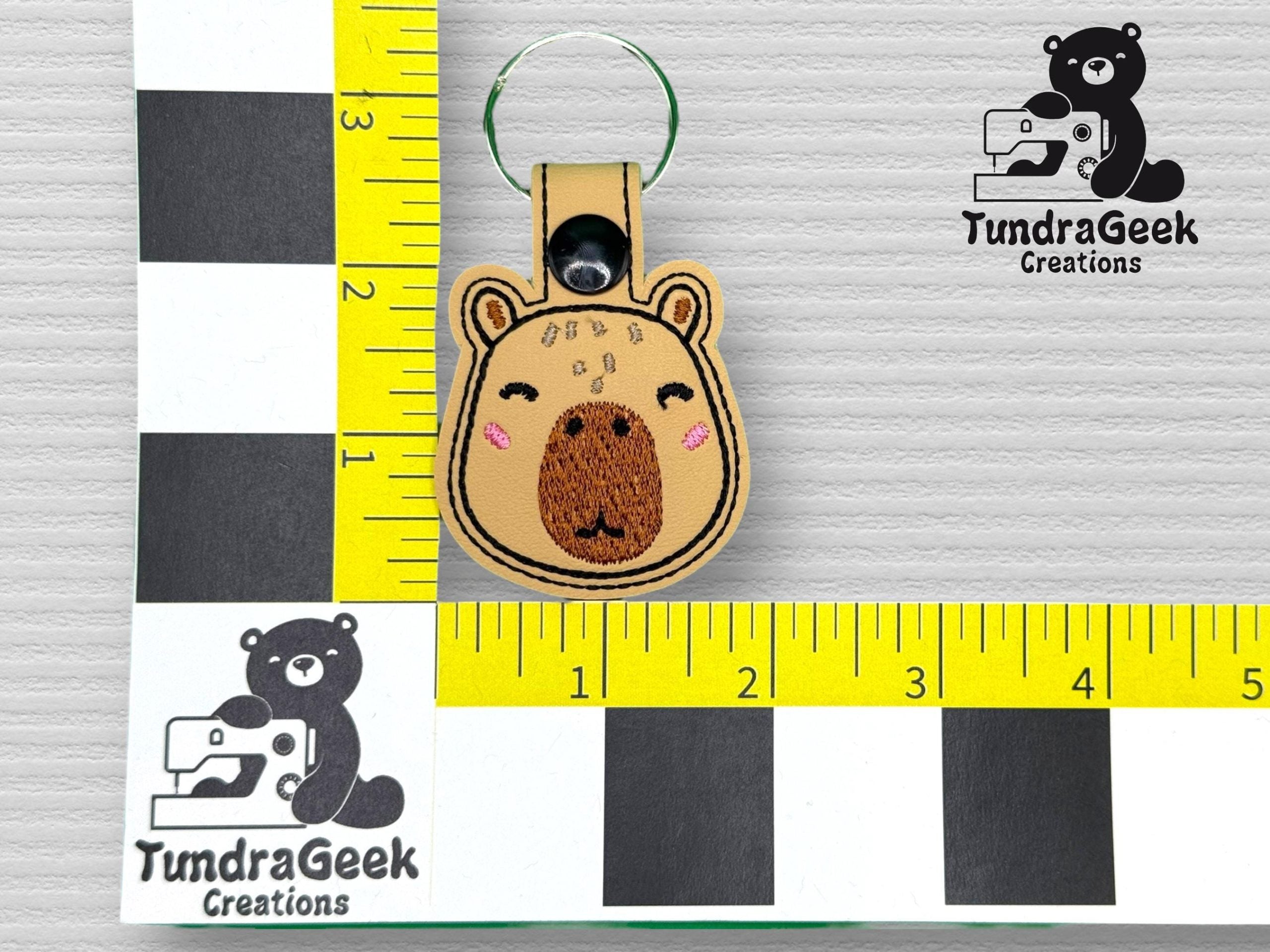 A bear-shaped embroidered keychain with a snap button lies on a measurement grid. TundraGeek Creations logo is visible in the top right corner.